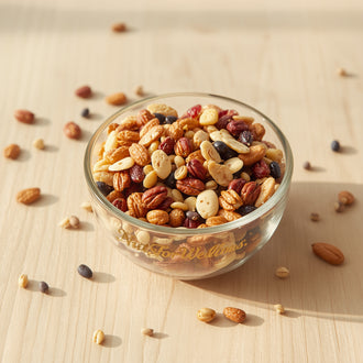 A glass bowl of NutsForWellness Healthy Trail Mix, featuring a vibrant blend of nuts, dried fruits, and seeds, sits on a light wooden surface with some pieces scattered nearby, illuminated by gentle natural light.