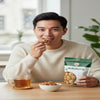 Man eating walnuts from a package with a bowl and cup of tea on a table.